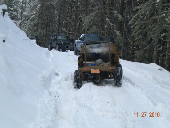 Several off-road vehicles driving on a snow-covered trail in a forest. The lead vehicle is a heavily modified, stripped-down utility vehicle. A blue pickup truck and a black Jeep are behind it