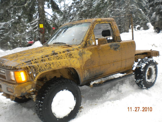 Yellow Toyota pickup truck covered in mud and snow, parked in a snowy wooded area. Date stamp reads 11.27.2010