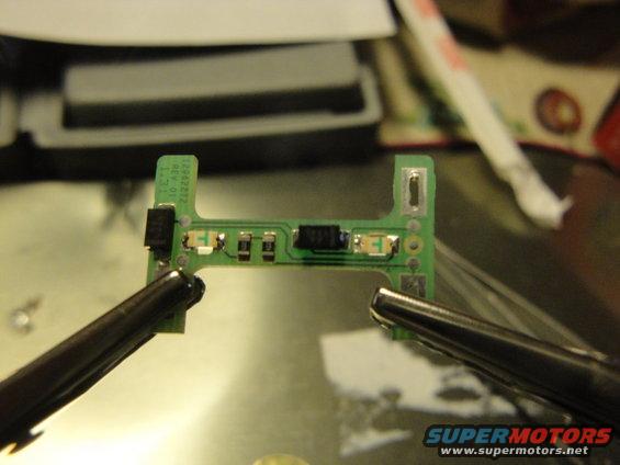 Close-up of a circuit board held by tweezers, with visible components and text