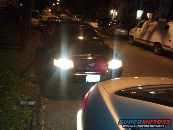 Front view of a 2003 Ford Crown Victoria parked on a street at night, headlights on. Another car is in the foreground