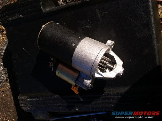 Starter motor with black housing and silver gear, resting on a black surface