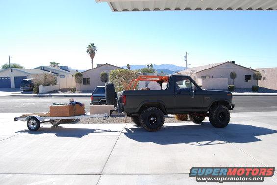 1982 Ford Bronco with aftermarket roll cage and spare tire carrier, towing a utility trailer