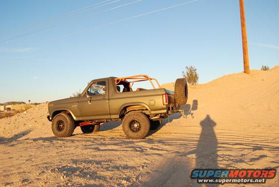 Olive green Ford Bronco with orange roll cage and spare tire on rear, driving on a dirt hill