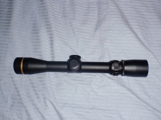 Black rifle scope with gold ring on the objective lens, lying on a striped fabric