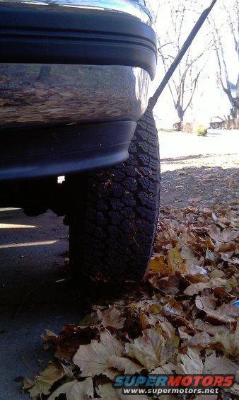 Close-up of a vehicle's rear tire and bumper, with fallen leaves on the ground