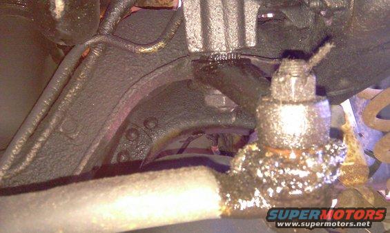 Close-up of a greasy ball joint and suspension arm, with a SuperMotors logo at the bottom right