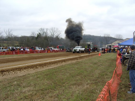 White pickup truck spewing black smoke during a pull contest, with spectators and other vehicles in the background