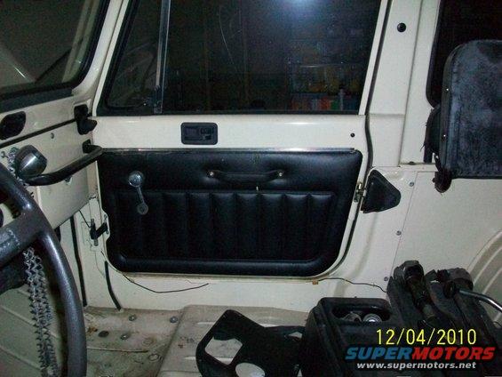 Interior shot of a Jeep's driver side door panel and dashboard area. Black padded door panel with a chrome crank handle