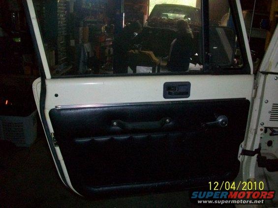White Jeep door with black vinyl panel, chrome handle, and window crank. Another vehicle is visible through the window