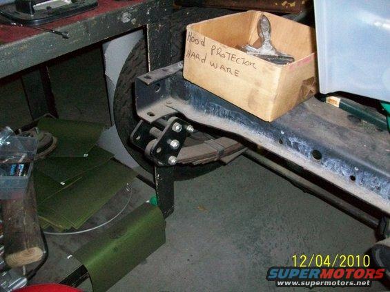 close-up of vehicle frame with leaf springs, a box labeled 'Hood PROTECTOR HARD WARE', and a tire