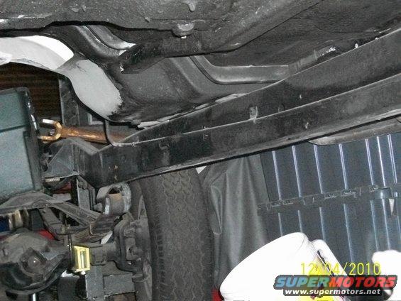 Underside view of a vehicle's frame, suspension, and fuel tank. A tire is visible