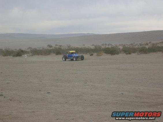 Blue dune buggy with yellow top driving on sand, desert landscape with hills and sparse vegetation