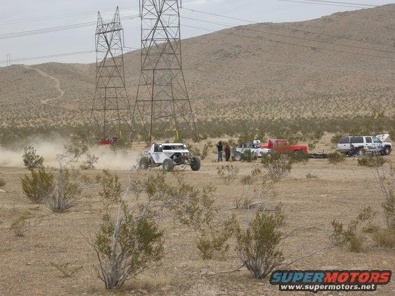 Off-road buggy kicking up dust near power lines, with several trucks parked in the desert