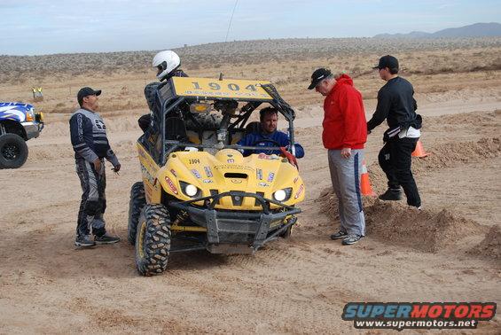 Yellow off-road buggy with people gathered around in a desert landscape. Another vehicle is visible to the left
