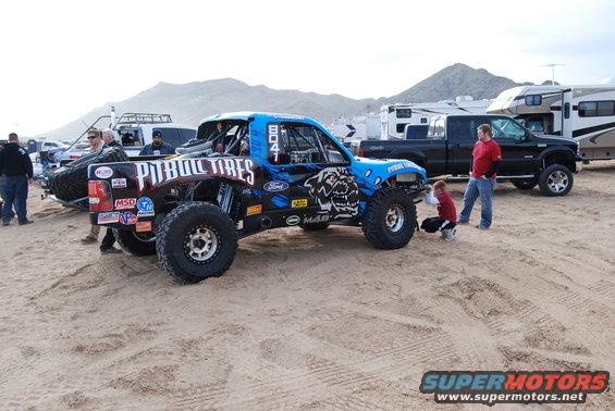 Blue off-road race truck with 'Pitbull Tires' on the side, parked in sand with other vehicles and RVs