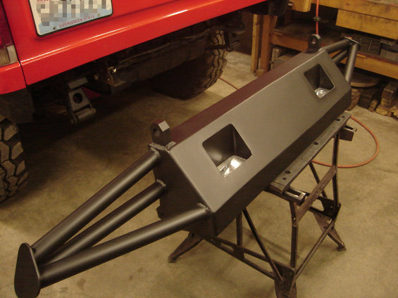 Custom steel rear bumper with dual light cutouts and triple tube protection, sitting on sawhorses. Red vehicle part visible to the left