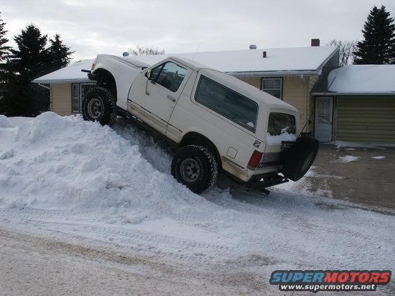 White SUV with lifted suspension and off-road tires climbing a large snow pile in front of a house