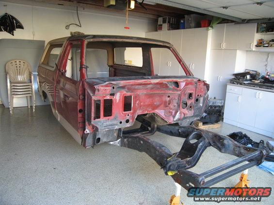 Red 1980s Ford Bronco body stripped down, chassis visible on stands in garage