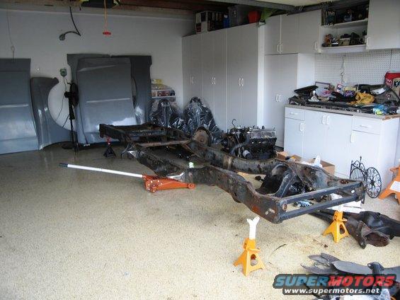 Vehicle frame on jack stands with engine nearby in a garage. Body panels are leaning against the wall
