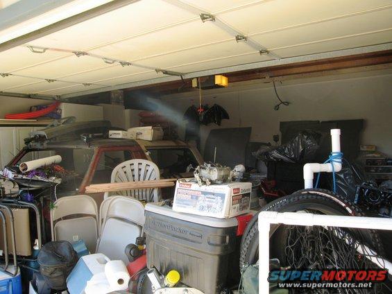 Garage interior cluttered with car parts, chairs, and storage bins