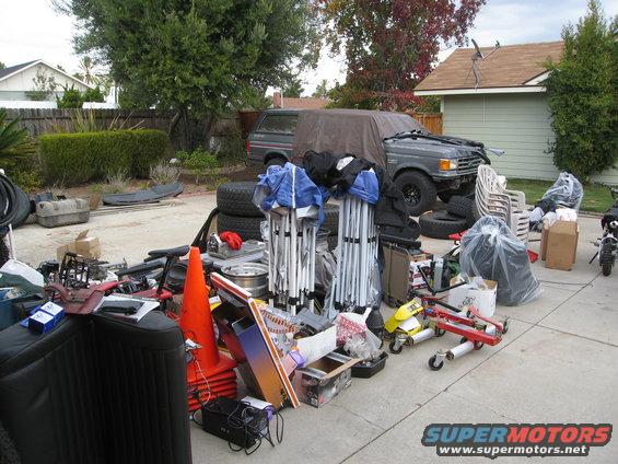 1987 Ford Bronco covered by a tarp, parked in a driveway with many items scattered around it