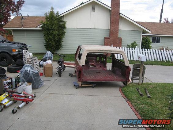 Ford Bronco body, stripped down, sitting on dollies in a driveway. Another vehicle is partially visible on the left