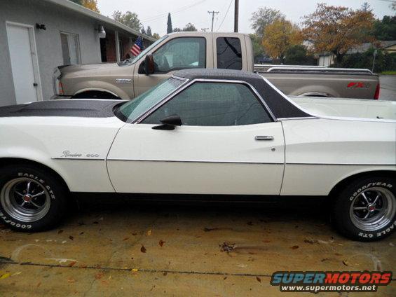 Side view of a white 1973 Ford Ranchero with a black hood and roof, parked next to a tan pickup truck