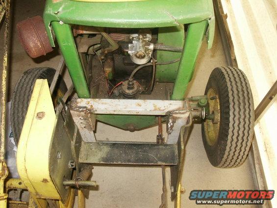 Top-down view of a green and yellow tractor's engine and front axle. Shows the carburetor, distributor, and wheels