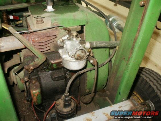 Green engine with carburetor, spark plug, and fuel filter visible; part of a tire is on the right