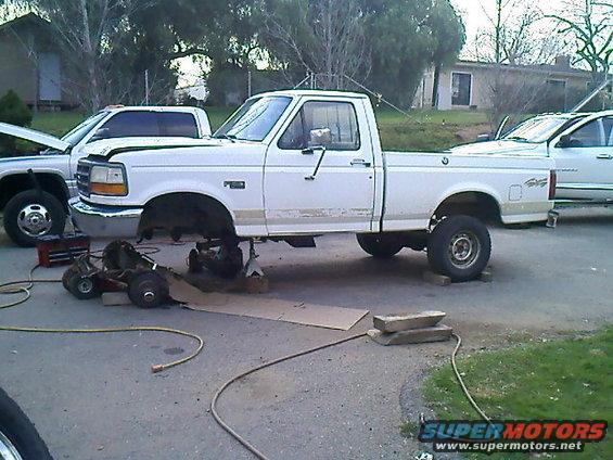 White pickup truck on jack stands with front end disassembled. Another truck and car are nearby