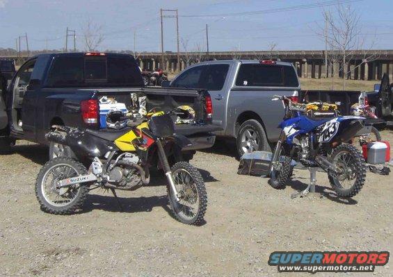 Dirt bikes and pickup trucks parked on a dirt lot with a bridge in the background