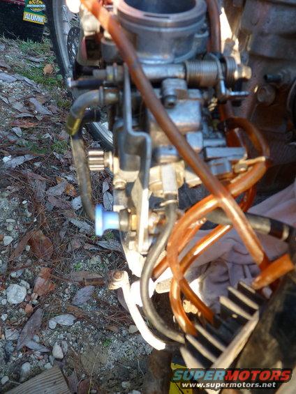 Motorcycle carburetor with orange fuel lines and a blue choke knob. A portion of a wheel and tire is visible