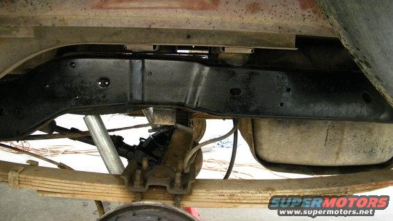 Underside of a vehicle showing a rear axle, leaf springs, and fuel tank. The frame appears to be from the 1980s