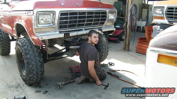 Man kneeling under lifted red Ford truck, working on front suspension. Another truck is visible to the right