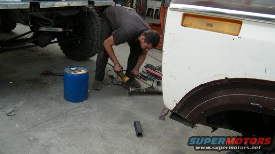 Man grinding on the rear fender area of a white SUV. Another vehicle's chassis and large tire are visible to the left
