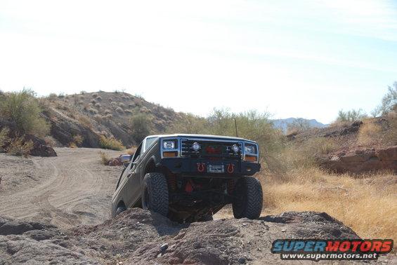 Dark green 1980s Ford Bronco off-roading on a rocky, desert trail with a modified bumper and large tires