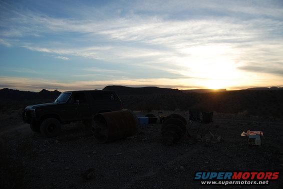 Dark Ford Bronco parked in a dusty, barren landscape at sunset with large metal drums and debris nearby