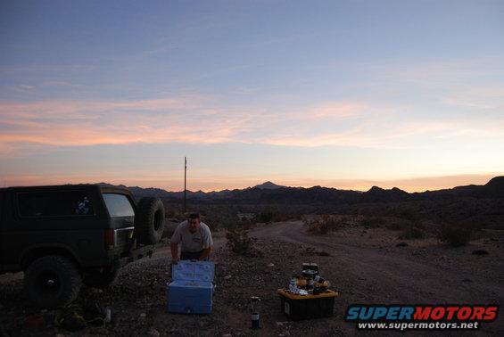 Man by cooler next to a dark-colored Ford Bronco with a spare tire on the back, desert landscape at dusk