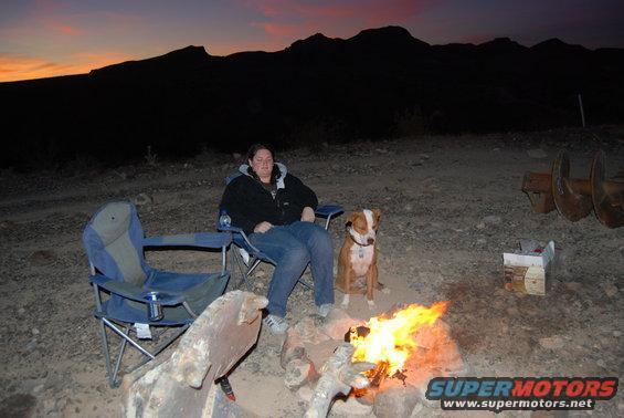 Camp scene at dusk with a woman and dog by a campfire, mountains in background