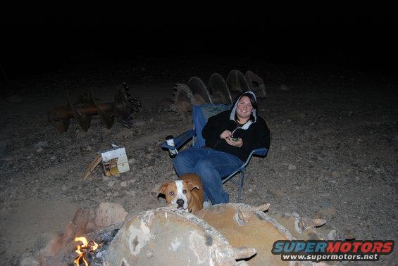 People and dogs sit around a campfire at night, with large auger bits in the background