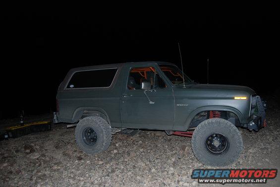 Dark green Ford Bronco with orange roll cage, lifted suspension, and large tires parked on gravel at night
