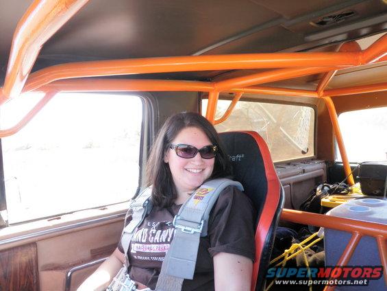 Woman wearing sunglasses and a racing harness sits in a vehicle with an orange roll cage