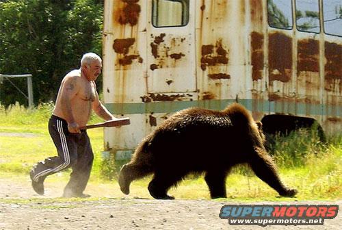 Man with a club runs after a bear next to a rusty, old bus or train car