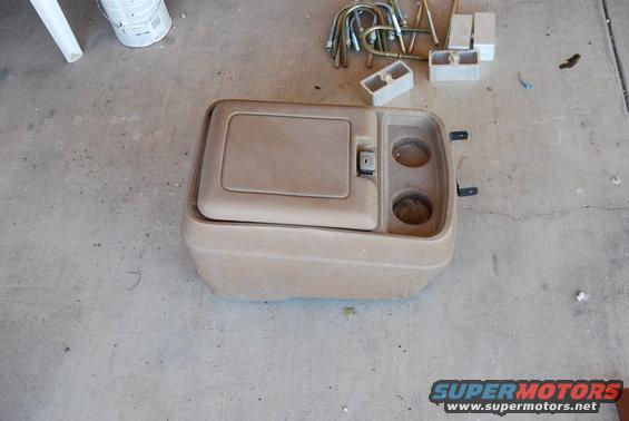 tan center console with a flip-top storage compartment and two cup holders, sitting on a concrete floor
