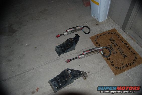 Two shock absorbers and three mounting brackets on a concrete surface next to a doormat