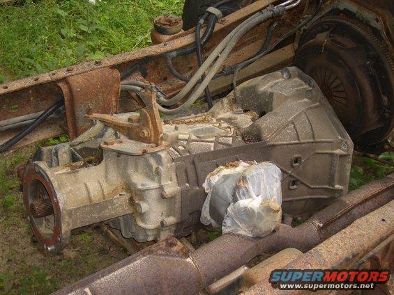 Rusty truck frame with a transmission assembly. A plastic-wrapped object rests on exhaust pipes