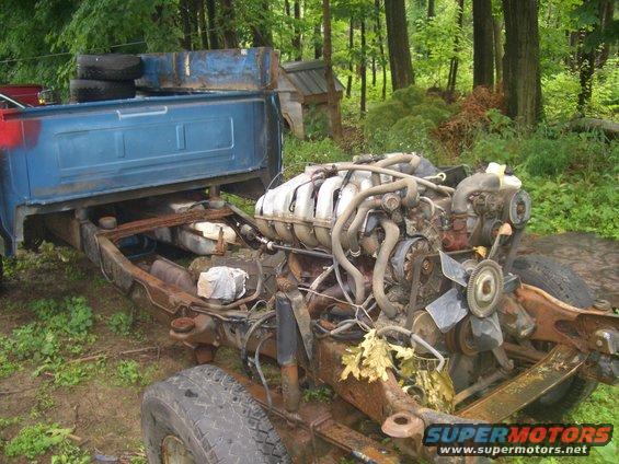 Blue truck bed and chassis with engine exposed, outdoors in a wooded area
