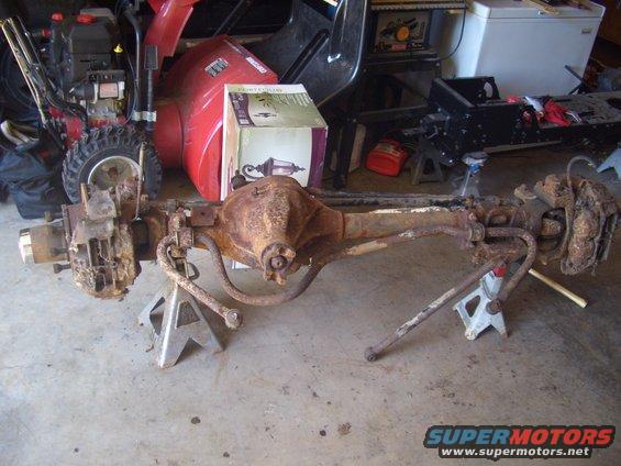 Rusty rear axle assembly on jack stands, with a snowblower and other items in the background