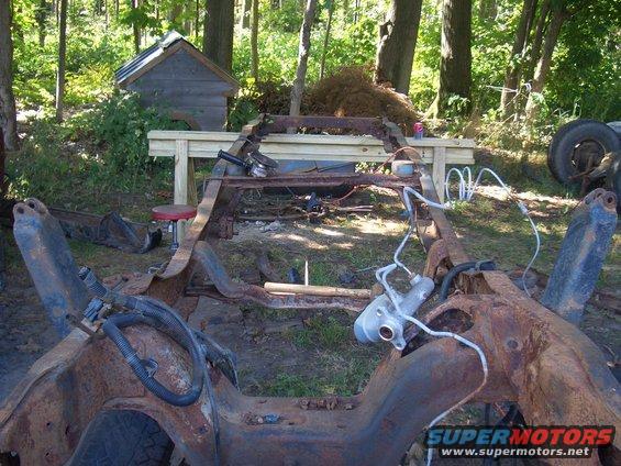 Rusted truck frame on grass, with trees and a small shed in background
