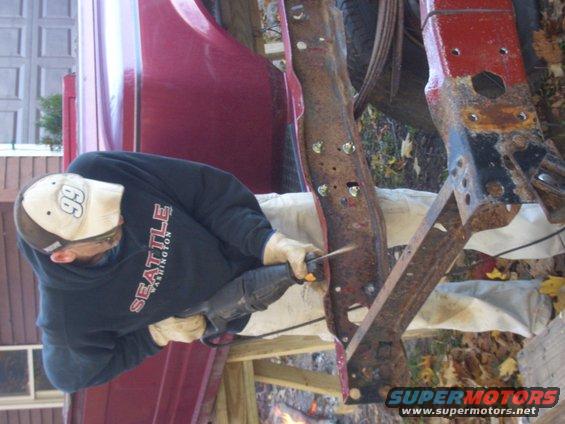 Man cutting rusty truck frame with reciprocating saw. Red body panel visible